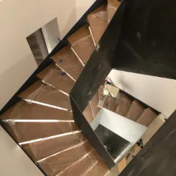 Wewnętrzne schody z drewnianymi stopniami zabezpieczonymi folią ochronną i czarną metalową balustradą, widok z góry w trakcie montażu.