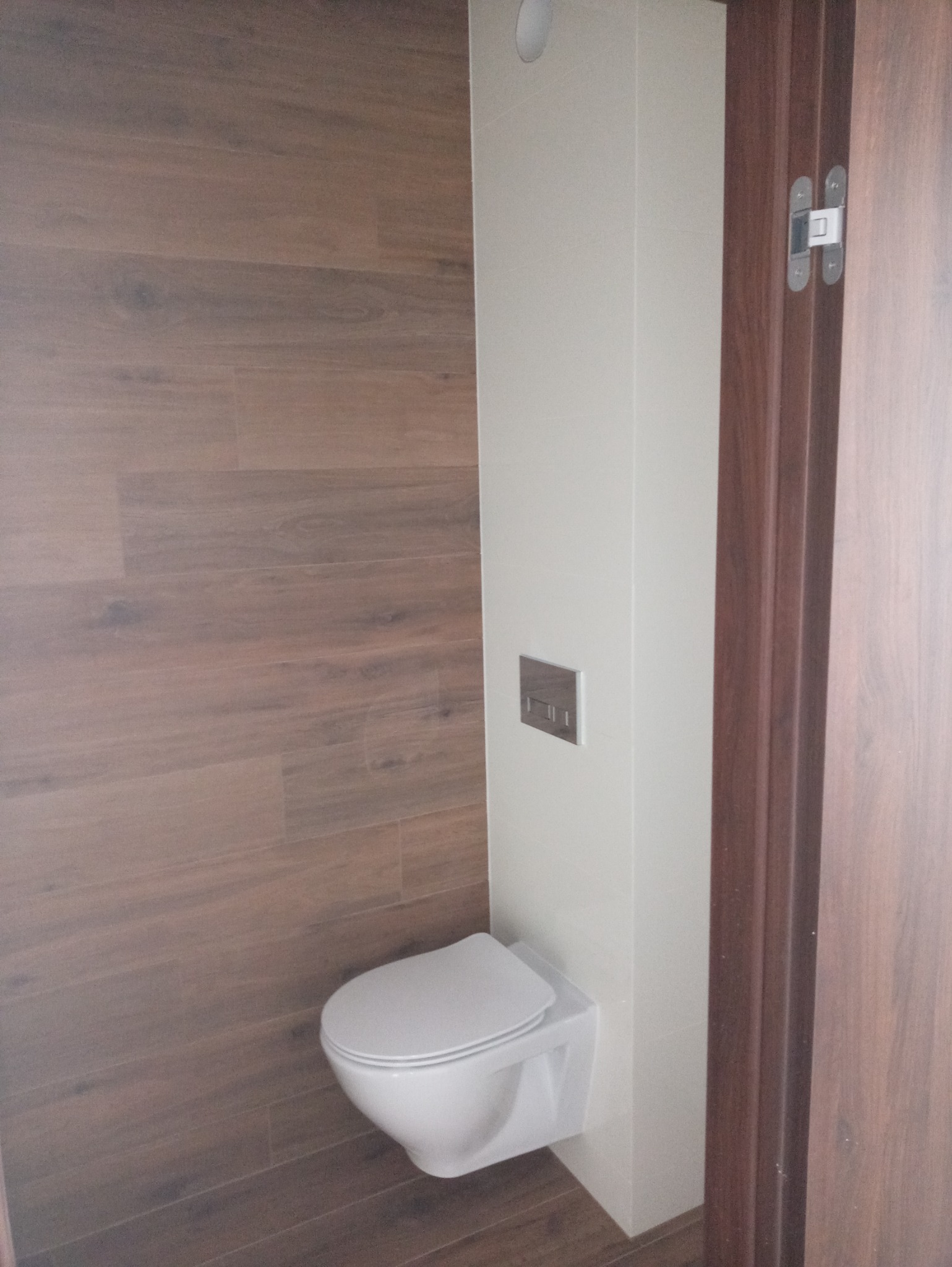 Nowoczesna toaleta podwieszana z chromowanym przyciskiem spłukiwania na tle ściany wyłożonej płytkami imitującymi drewno i białych płytek, widoczna część brązowej ościeżnicy.