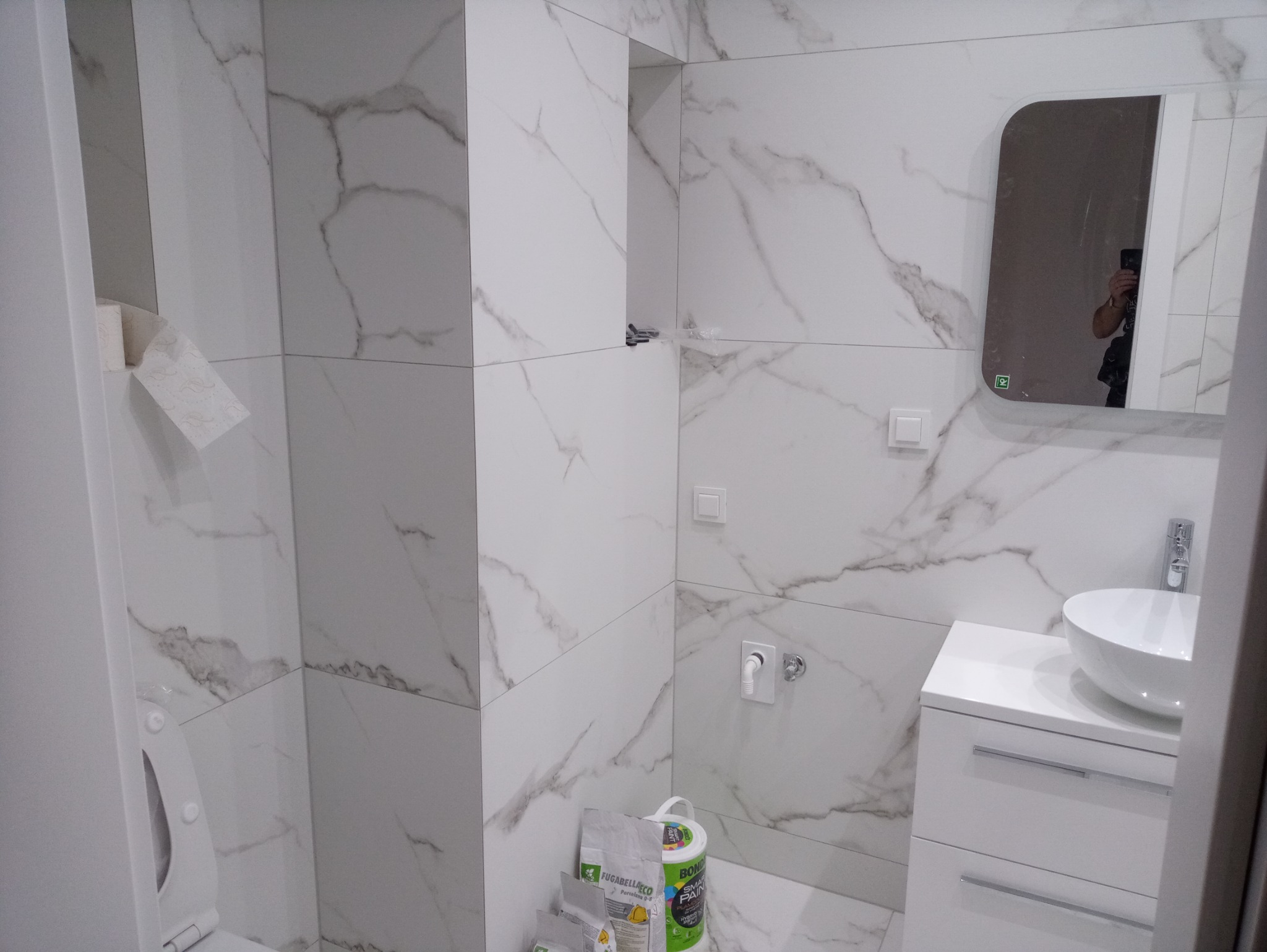 Wykończona łazienka z białymi płytkami imitującymi marmur, umywalką nablatową i lustrem z zaokrąglonymi rogami; widoczne worki z zaprawą i wiadro farby.