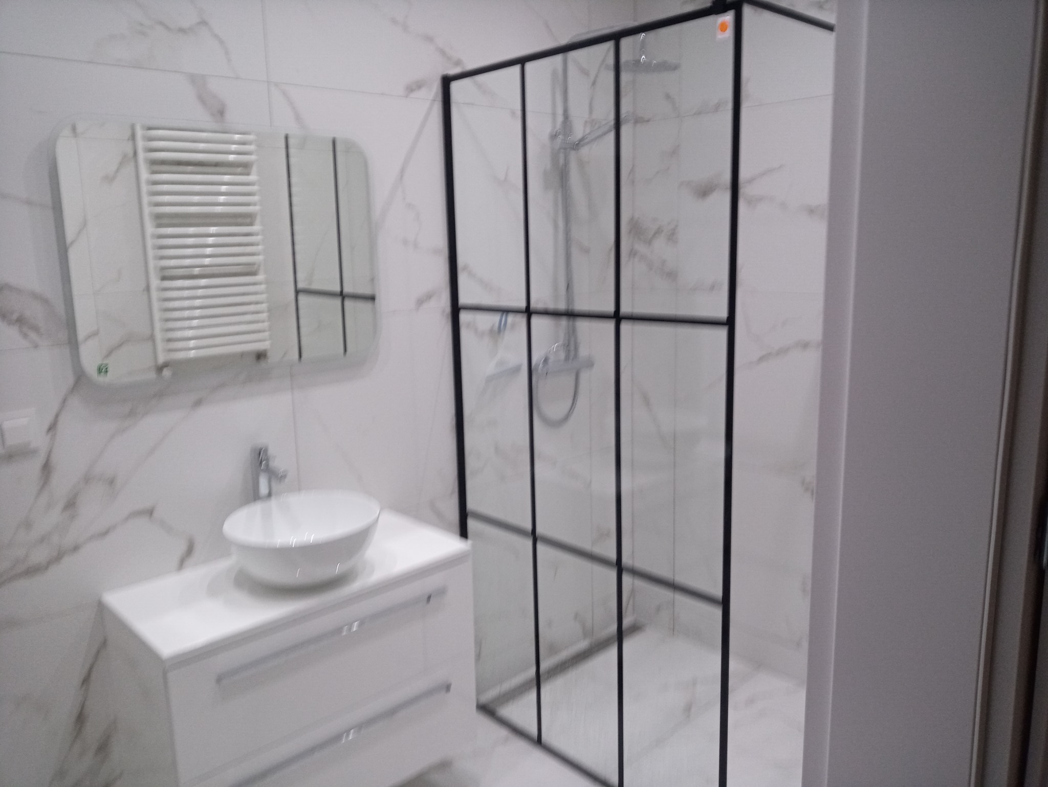Nowoczesna łazienka z białą umywalką nablatową, grzejnikiem drabinkowym w lustrze i kabiną prysznicową z czarnymi profilami na tle marmurowych płytek.