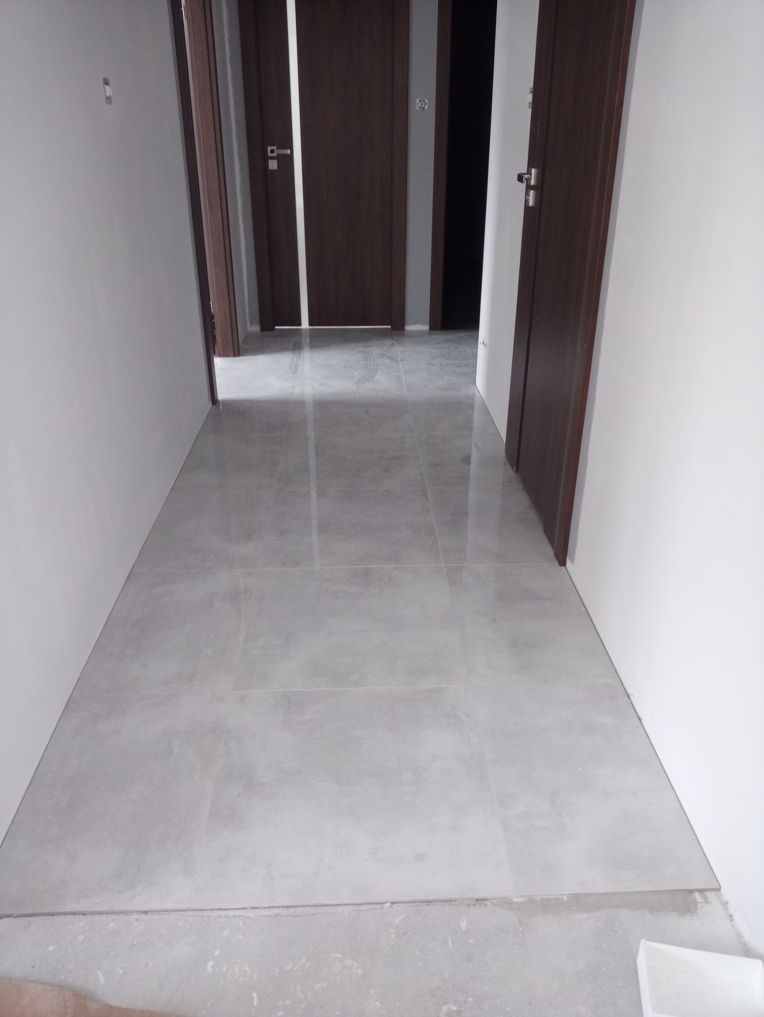 Korytarz z ułożoną szarą, matową podłogą z płytek ceramicznych o dużym formacie, widoczne białe ściany i ciemne, drewniane drzwi wewnętrzne.