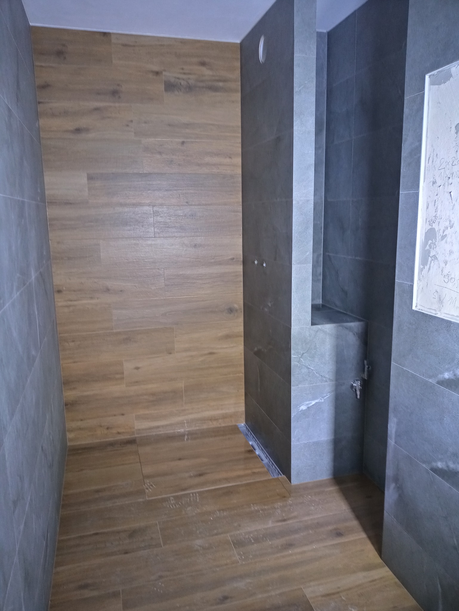 Wykończona łazienka z drewnopodobnymi płytkami na podłodze i ścianie oraz szarą kabiną prysznicową typu walk-in z odpływem liniowym; widoczne otwory na baterię.