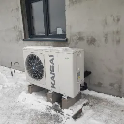 Zewnętrzna jednostka pompy ciepła Kaisai zamontowana na betonowych bloczkach, w zimowej scenerii, z surowym tłem ściany budynku i śladami w śniegu.