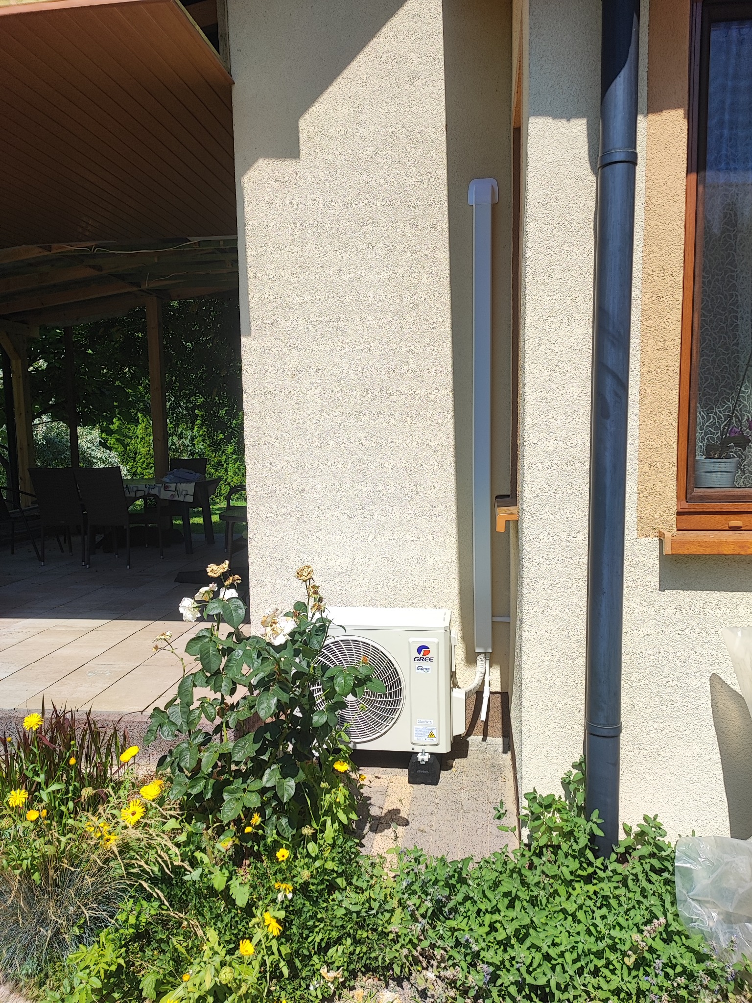 Biała jednostka zewnętrzna klimatyzacji marki Gree zamontowana na elewacji domu, obok rynny spustowej, z widocznym fragmentem ogrodu z kwiatami i zadaszenia tarasu.
