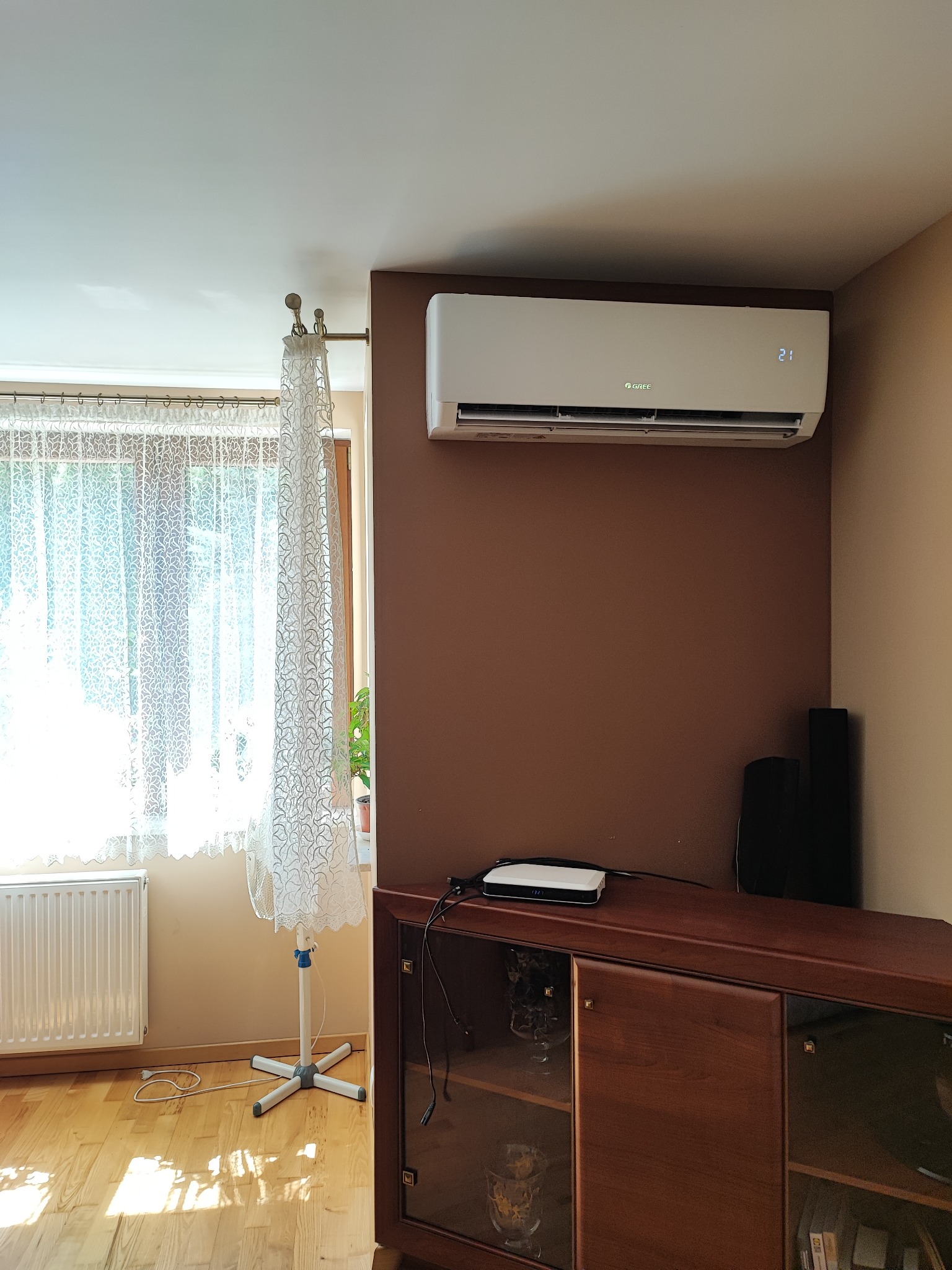 Biały klimatyzator Gree zamontowany na brązowej ścianie w pokoju z drewnianą komodą, firanką i grzejnikiem.