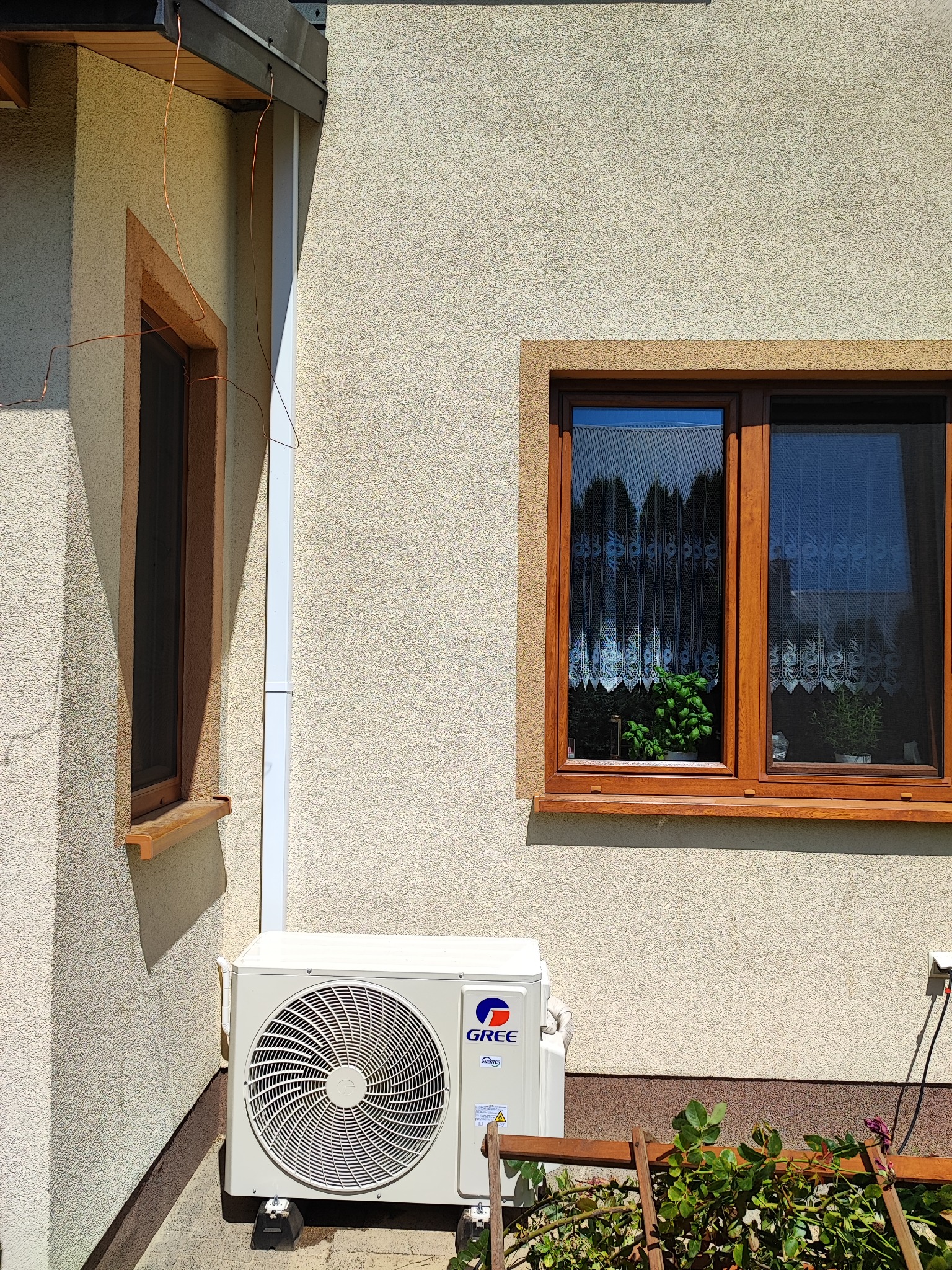 Biała jednostka zewnętrzna klimatyzatora marki Gree zamontowana na elewacji domu, widoczna spod okna z drewnianą ramą, z fragmentem drewnianej podpory dla roślin w dolnej części kadru.
