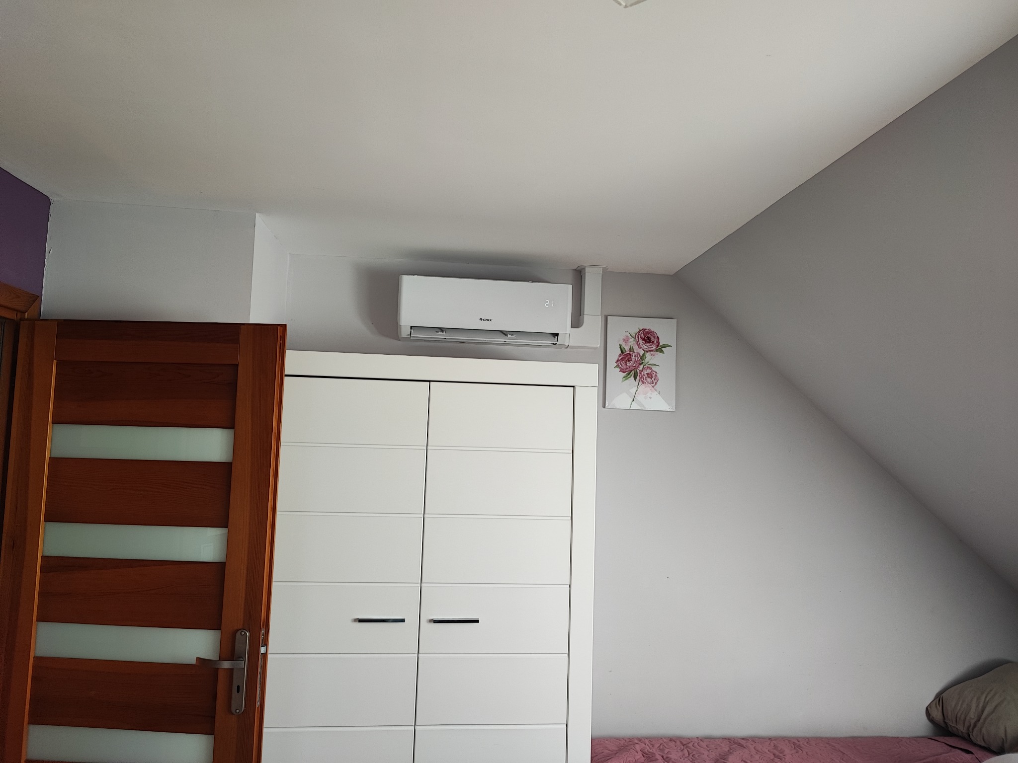 Biała jednostka klimatyzacyjna Gree zamontowana nad szafą wnękową w pokoju na poddaszu, obok obrazka z różami.