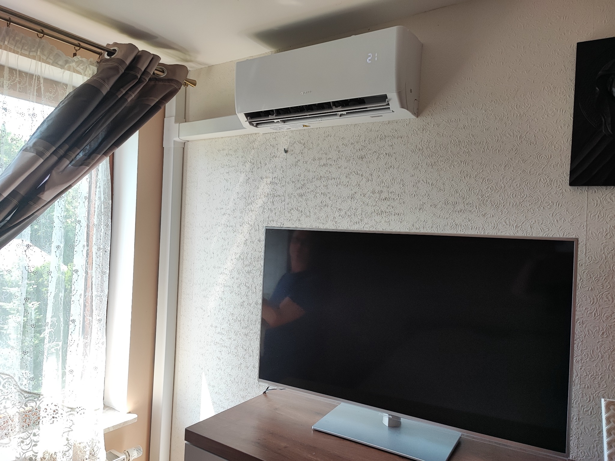 Biały klimatyzator marki Gree zamontowany na ścianie nad telewizorem w pokoju z wzorzystą tapetą i zasłoniętym oknem.