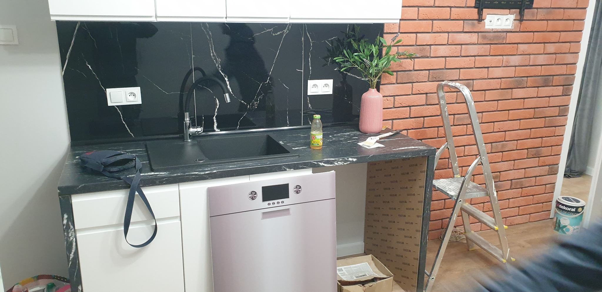 Kuchnia w trakcie remontu: czarny marmur na ścianie, zlew i blat, ceglana ściana, drabina malarska i niedokończona zabudowa pod blatem.