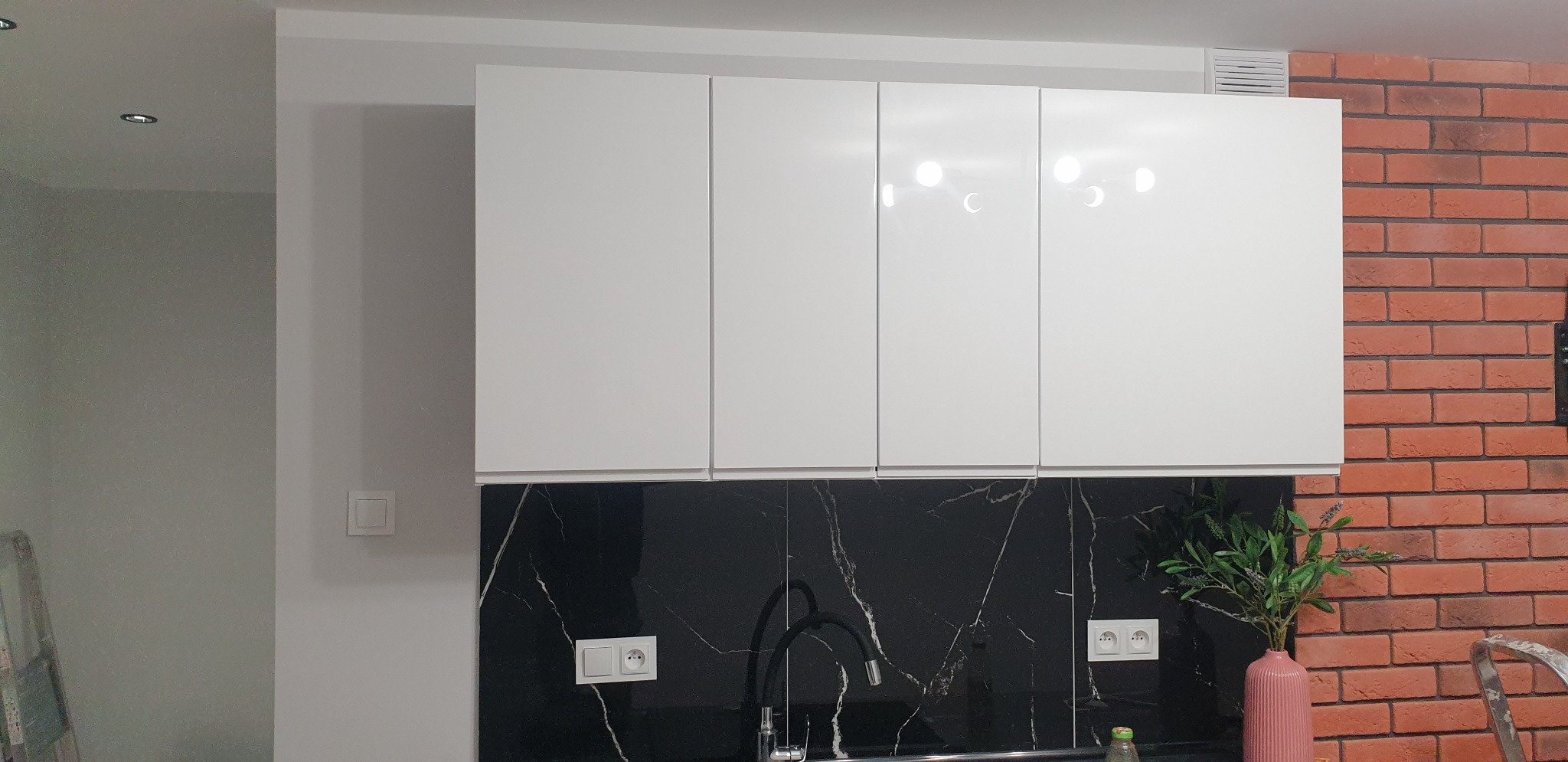 Nowoczesna kuchnia z białymi, błyszczącymi szafkami wiszącymi, czarnym blatem imitującym marmur z widoczną baterią i gniazdkami elektrycznymi, oraz ścianą z czerwonej cegły, uzupełniona różowym...