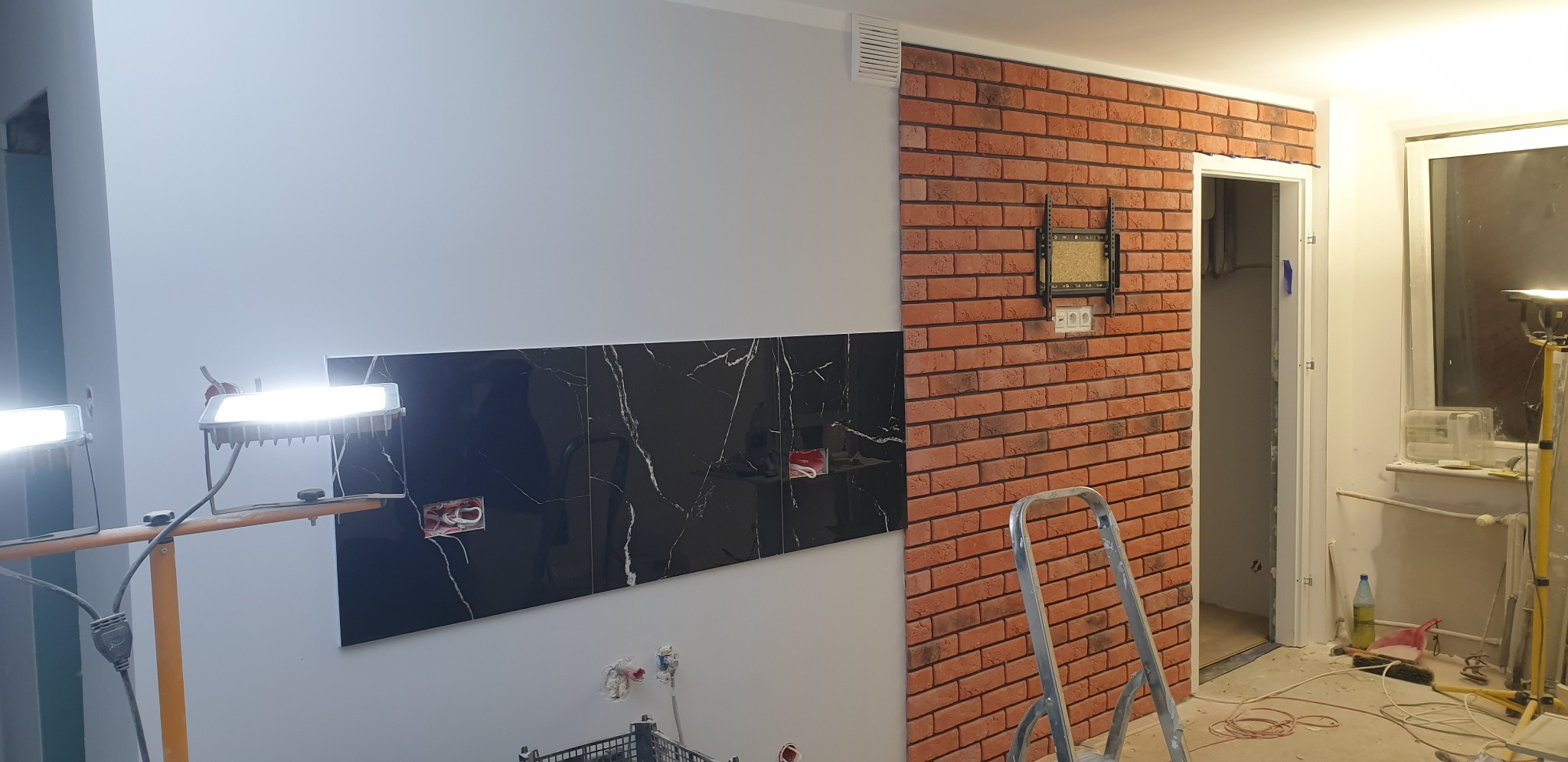 Częściowo wykończona ściana z czarnych płytek imitujących marmur i czerwonej cegły, widoczne puszki elektryczne, drabina i oświetlenie budowlane.