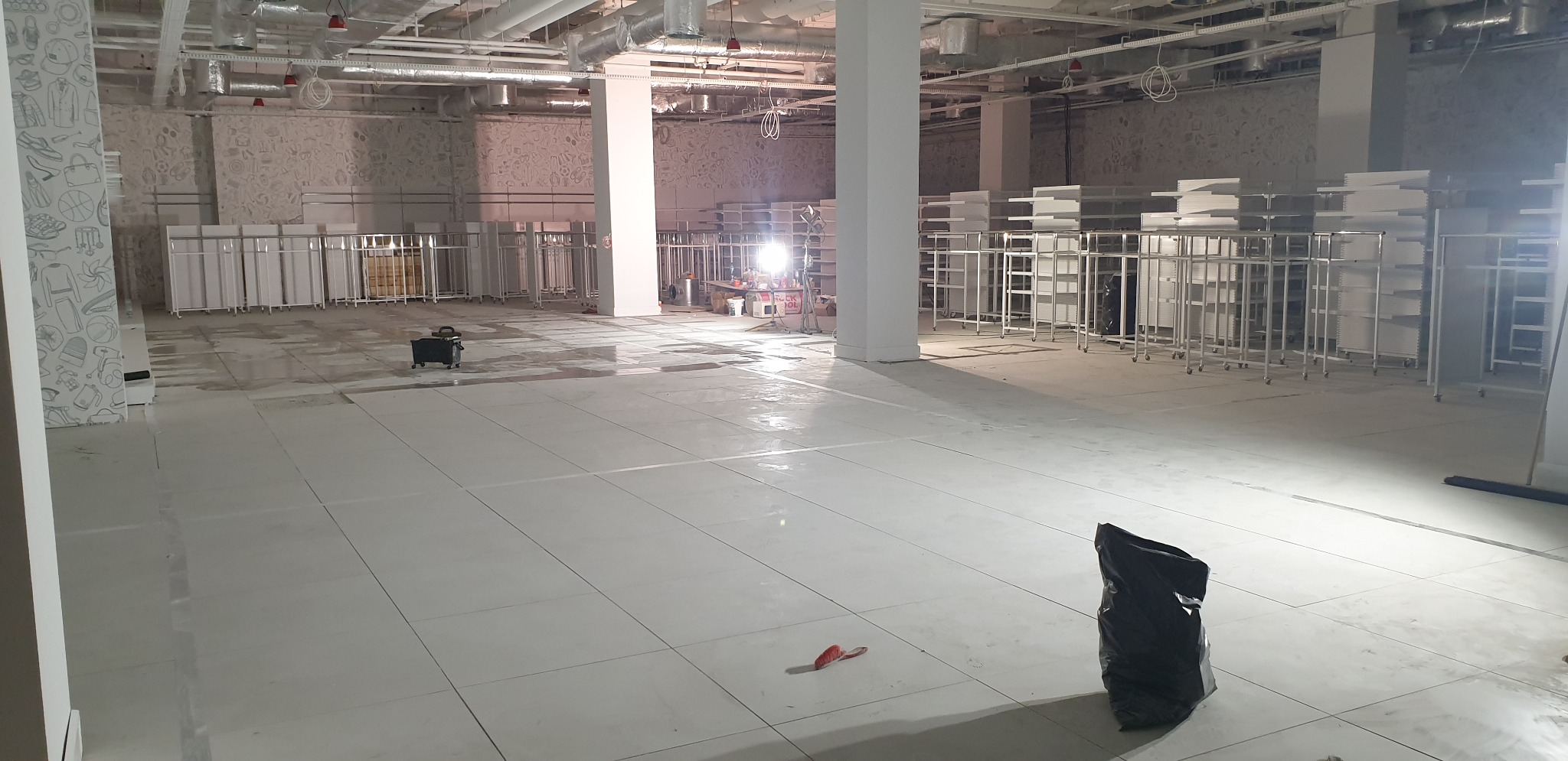 Wnętrze sklepu w trakcie remontu, widoczne białe płytki podłogowe, metalowe regały na kółkach i narzędzia budowlane, surowe ściany z dekoracyjnym wzorem.