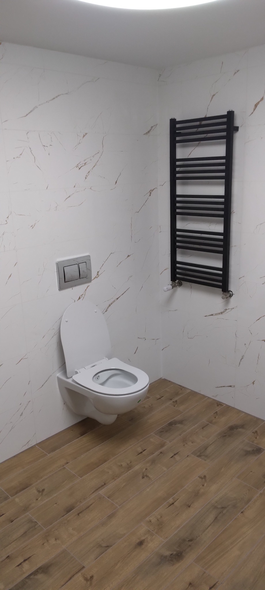 Nowoczesna łazienka z wiszącą toaletą, marmurowymi płytkami ściennymi z delikatnym żyłkowaniem i drewnopodobną podłogą, uzupełniona czarnym, drabinkowym grzejnikiem.