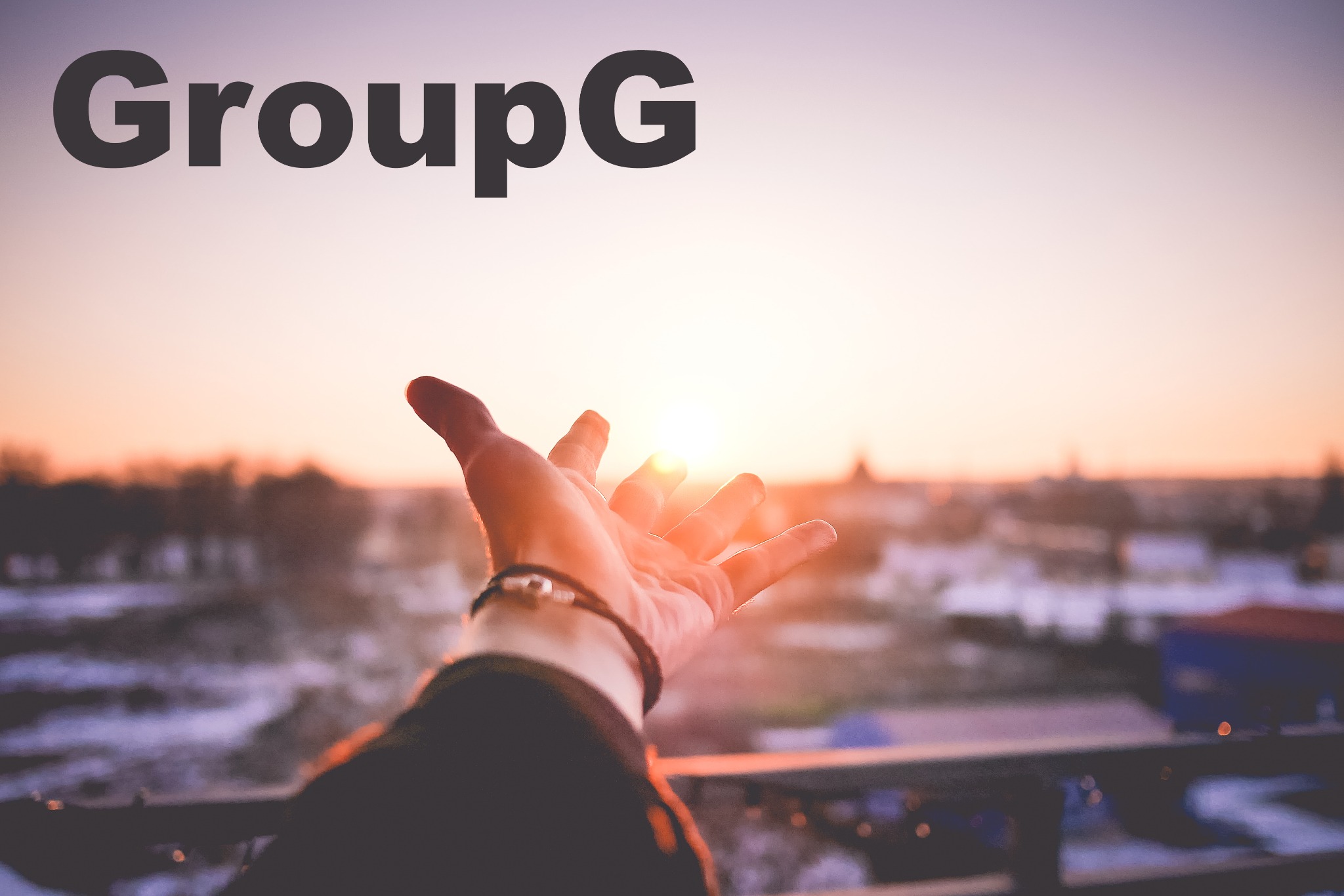Dłoń z bransoletką wyciągnięta w stronę zachodzącego słońca nad miastem, z widocznym napisem 'GroupG' u góry.