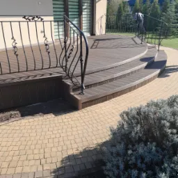 Zewnętrzny taras z desek kompozytowych w odcieniach brązu, z ozdobną, metalową balustradą i schodami, prowadzący do domu z dużymi oknami, otoczony brukowaną ścieżką i zielenią.