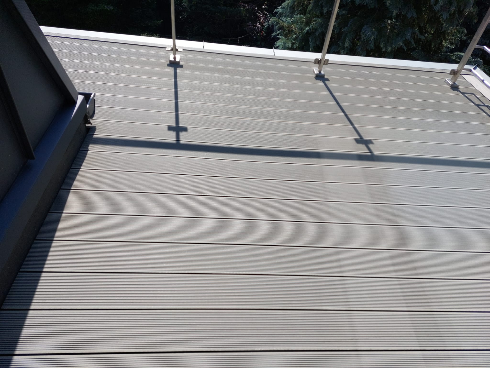 Szare deski tarasowe z widocznymi mocowaniami balustrady i fragmentem orynnowania, ułożone poziomo, z drzewami w tle.