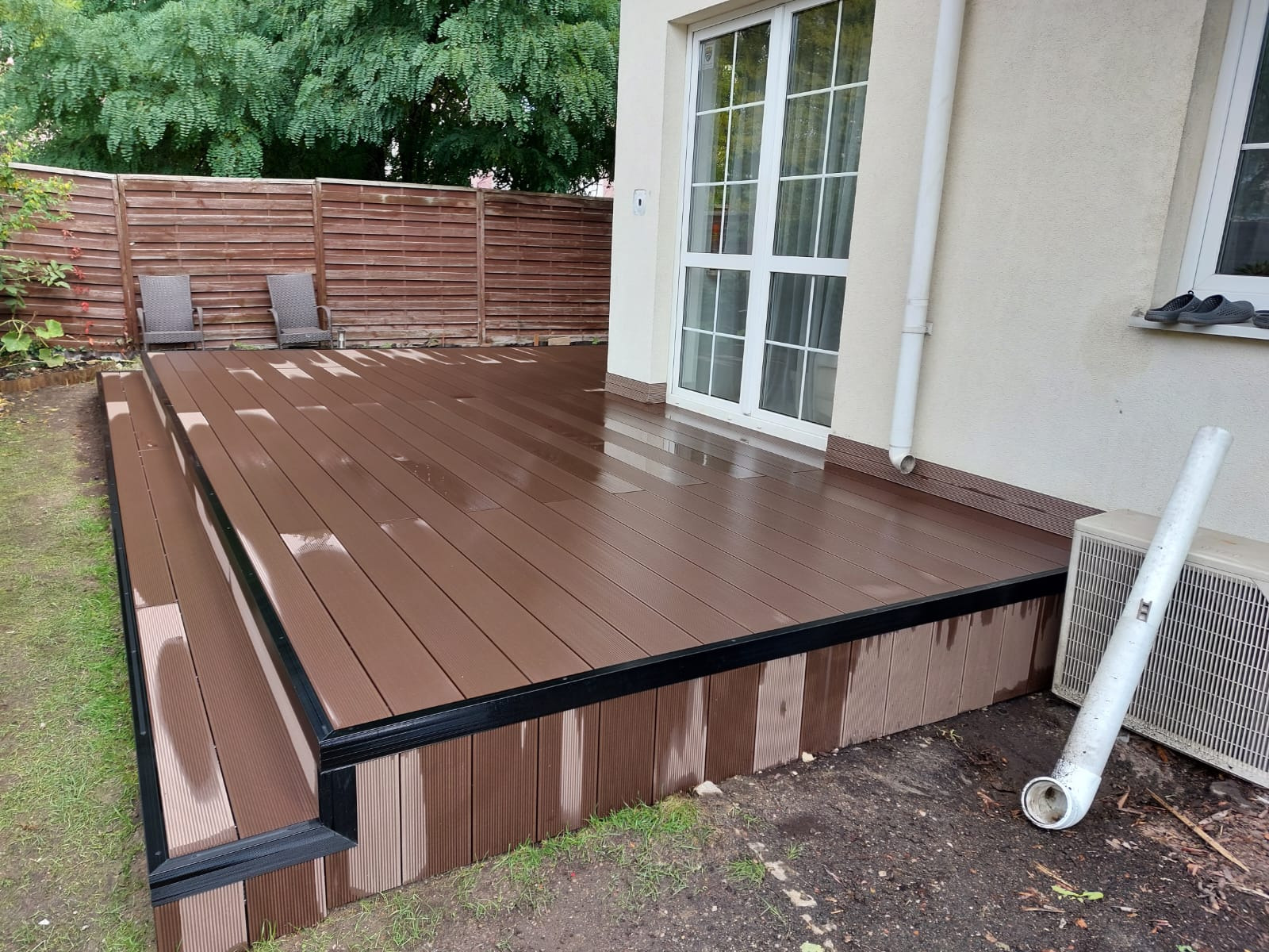 Nowoczesny taras z brązowych desek kompozytowych z dwoma stopniami, otoczony zielenią i drewnianym ogrodzeniem, widok na białe drzwi balkonowe i elementy instalacji na elewacji.