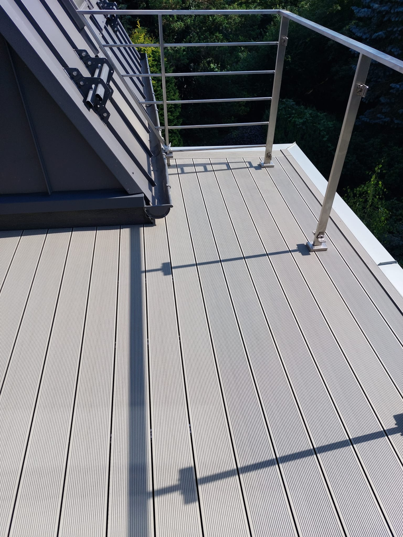 Jasnoszary taras kompozytowy z metalową balustradą, widoczne mocowania balustrady i cień słupka na deskach.