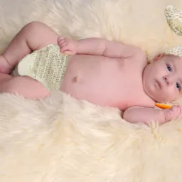 Niemowlę leżące na futrzanym dywanie, ubrane w dziergany kapelusik z uszami królika i pieluszkę, trzymające w dłoni pluszową marchewkę.