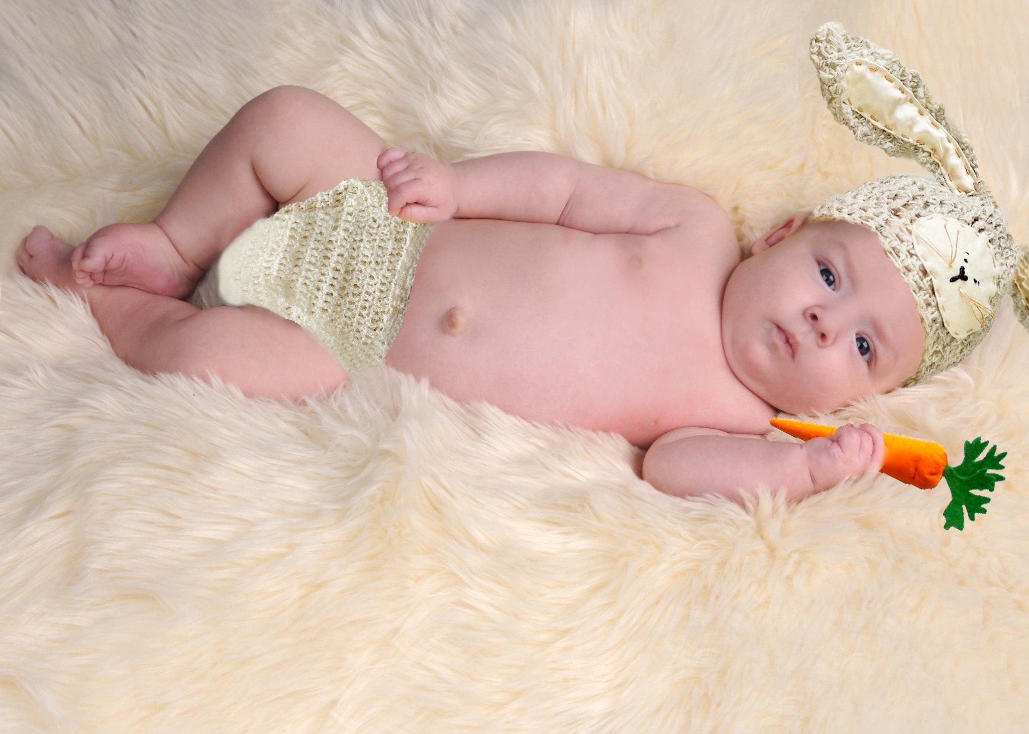 Niemowlę leżące na futrzanym dywanie, ubrane w dziergany kapelusik z uszami królika i pieluszkę, trzymające w dłoni pluszową marchewkę.