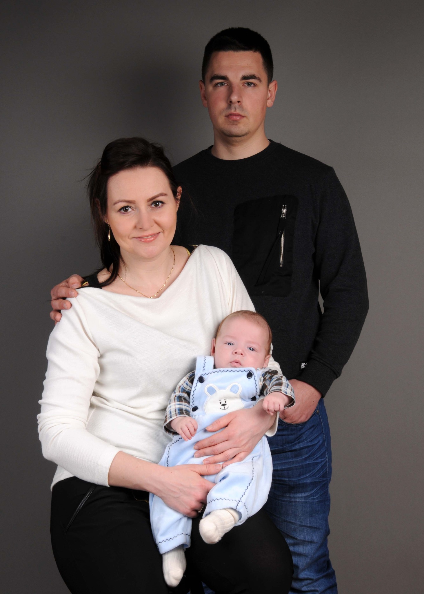 Rodzinne portretowe ujęcie: matka trzyma niemowlę ubrane w niebieski kombinezon z aplikacją misia, ojciec stoi za nimi na szarym tle.