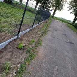 Nowo postawione, czarne ogrodzenie panelowe z podmur&oacute;wką betonową, widoczne od strony drogi, z nieuporządkowanym pasem ziemi wzdłuż ogrodzenia.