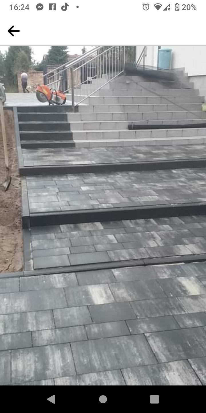 Ułożona szara kostka brukowa prowadząca do schodów z metalową barierką; w tle mężczyzna i piła do cięcia kostki.