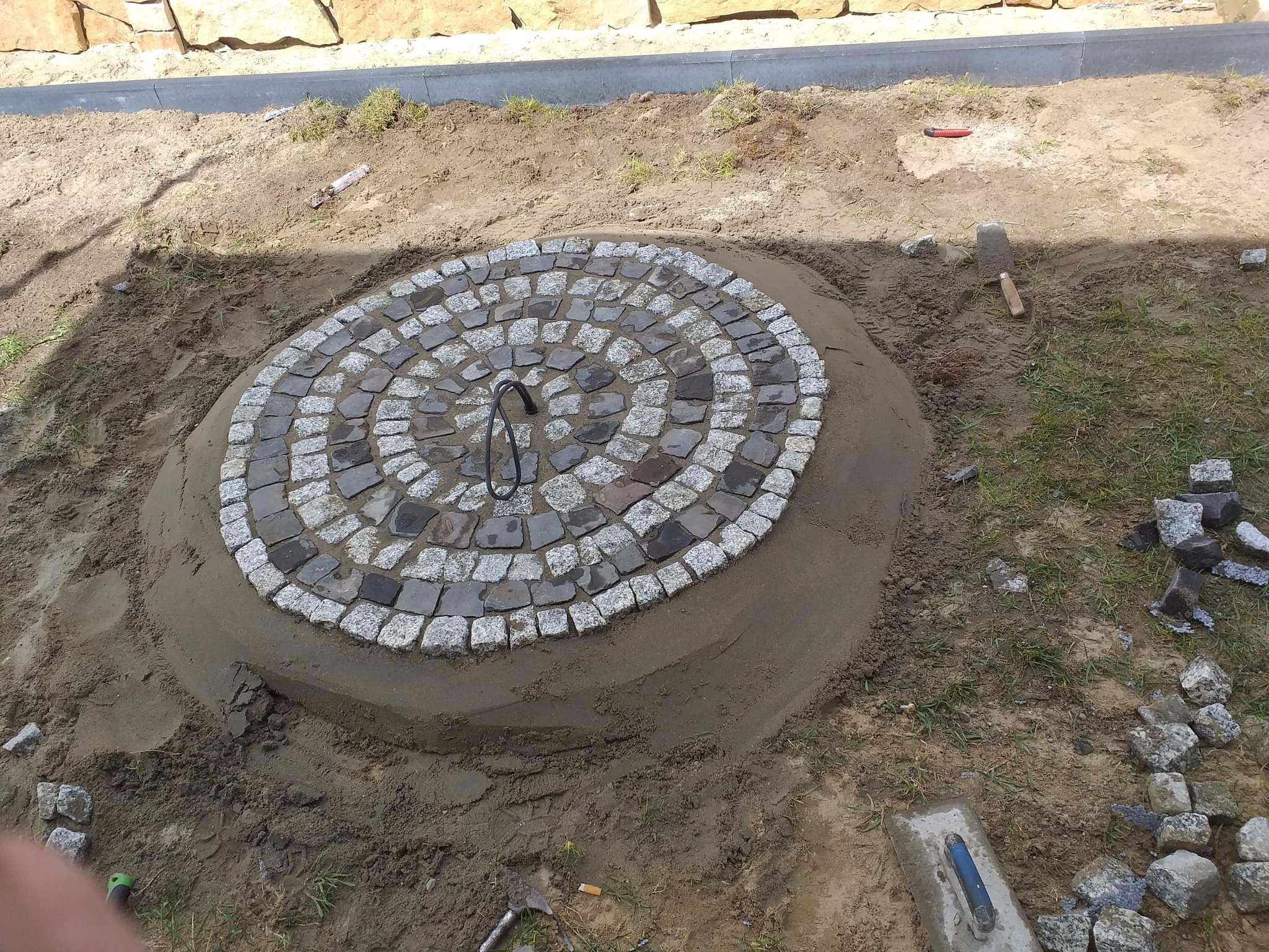 Okrągły wzór z kostki granitowej w trakcie układania, otoczony piaskiem i ziemią, z widocznymi narzędziami brukarskimi.