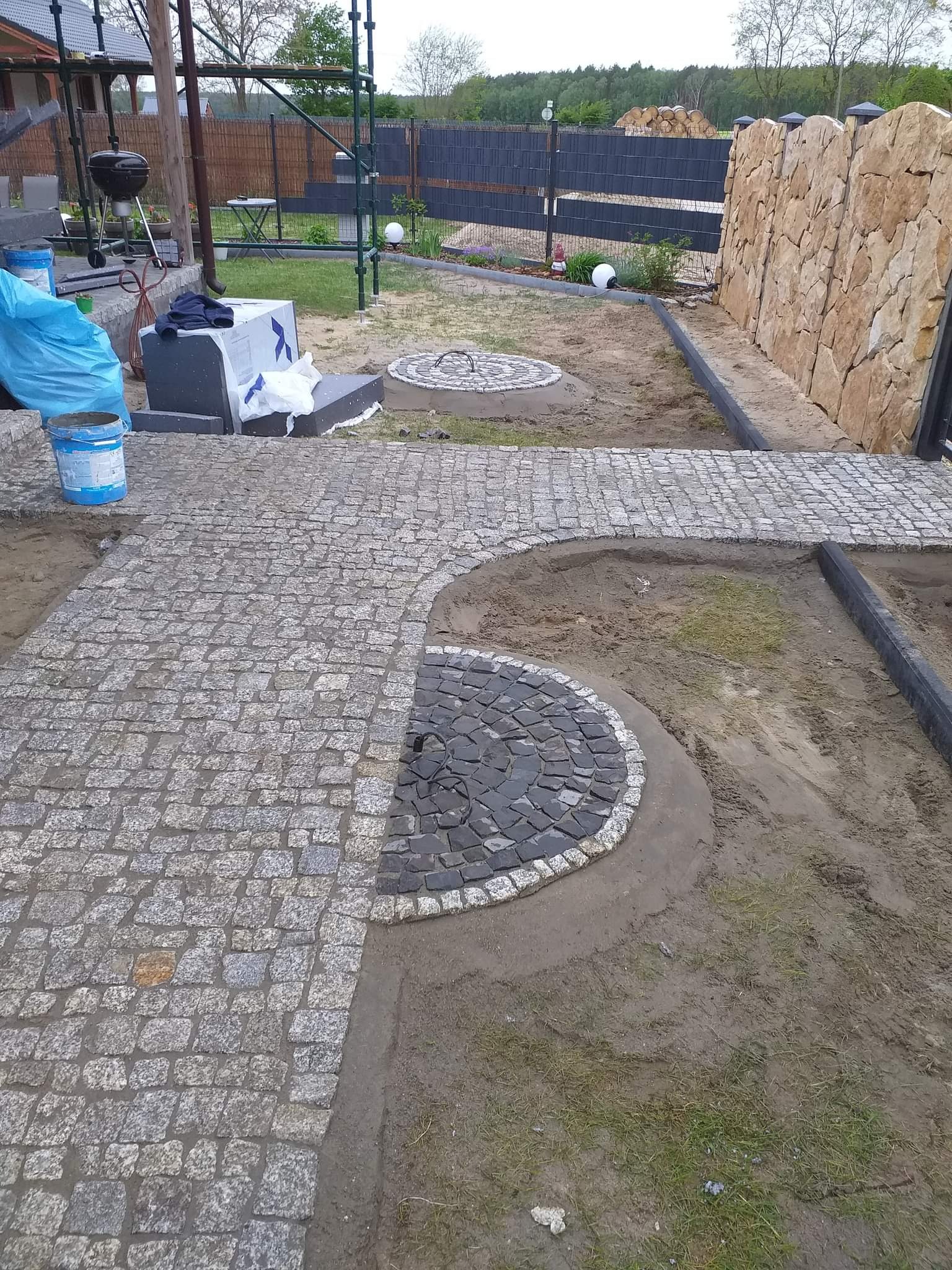 Fragment ścieżki z szarej kostki granitowej przechodzącej w ciemny okrąg z obwódką, w tle trawnik, grill i ogrodzenie.