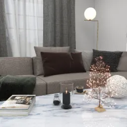 Jasny salon z szarym narożnikiem, marmurowym stolikiem kawowym, dekoracyjnym drzewkiem z lampkami LED, świecą w szklanym lampionie oraz filiżanką kawy.