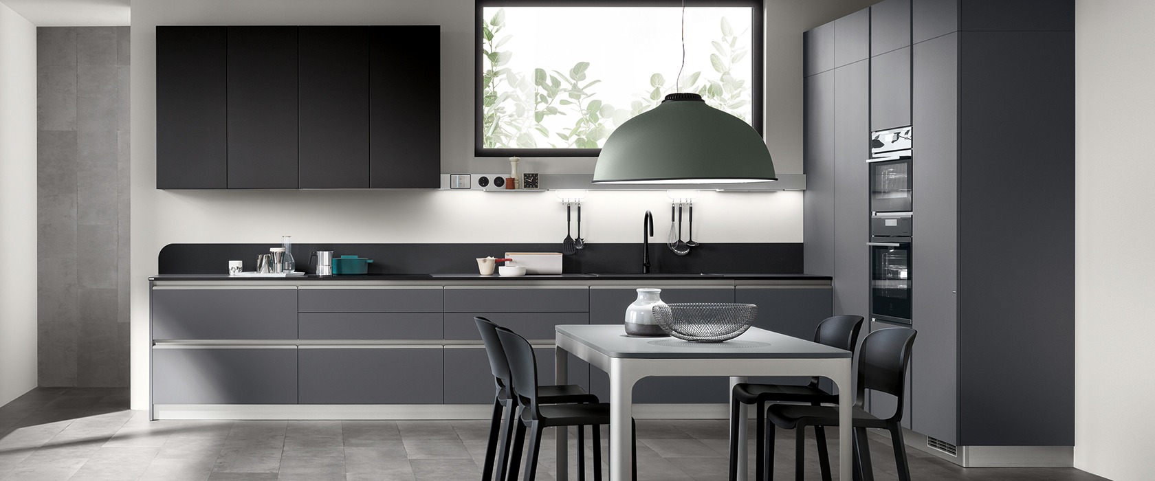 Nowoczesna kuchnia w odcieniach szarości z czarnymi akcentami, oświetlona dużym, zielonym kloszem lampy wiszącej nad stołem z designerskimi krzesłami.