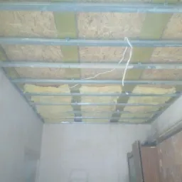 Montaż izolacji z wełny mineralnej w metalowym stelażu sufitu podwieszanego, widoczne przewody elektryczne i płyty OSB.