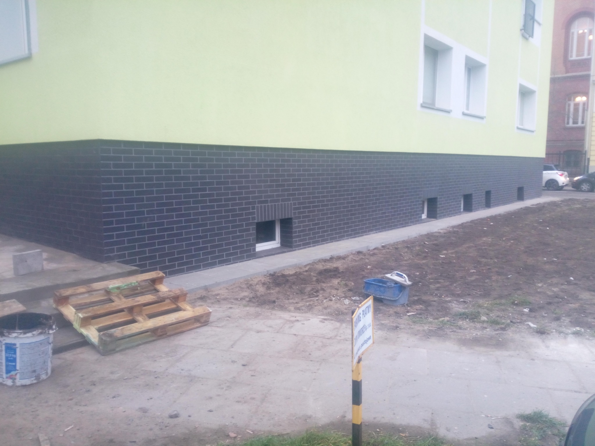 Elewacja budynku z wykończoną cokołową częścią z ciemnej cegły klinkierowej, widoczne paleta, wiadro i narzędzia murarskie, teren wokół w trakcie prac wykończeniowych.