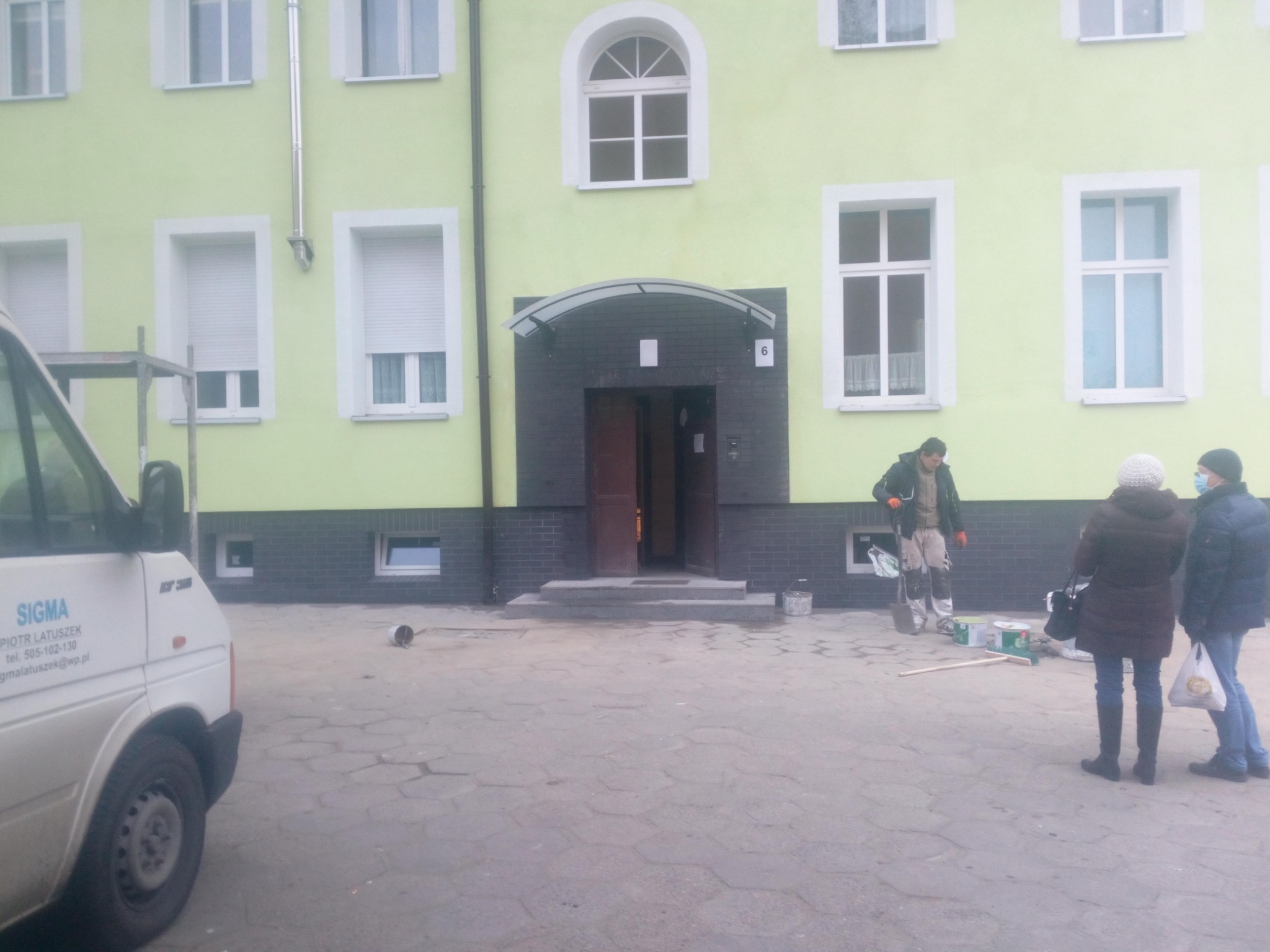 Pracownik w trakcie malowania elewacji budynku o jasnozielonym kolorze, widoczne puszki z farbą, obok stoją dwie osoby obserwujące prace, na pierwszym planie zaparkowany biały bus z logo firmy.