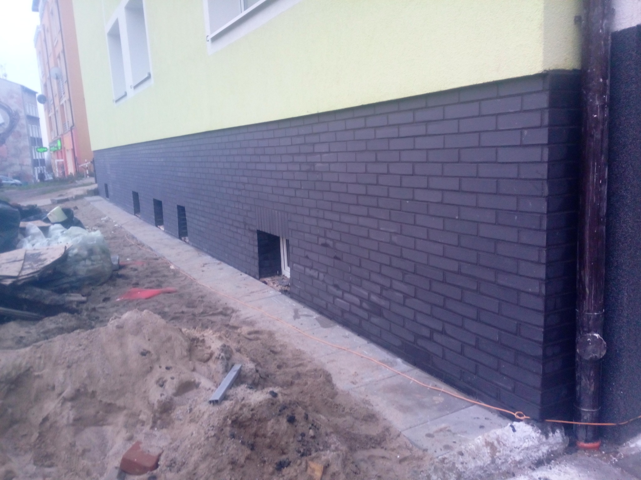 Wykończenie elewacji budynku z ciemnej cegły klinkierowej na fundamencie, widoczne otwory wentylacyjne, teren wokół w trakcie prac ziemnych, piasek i resztki materiałów budowlanych.