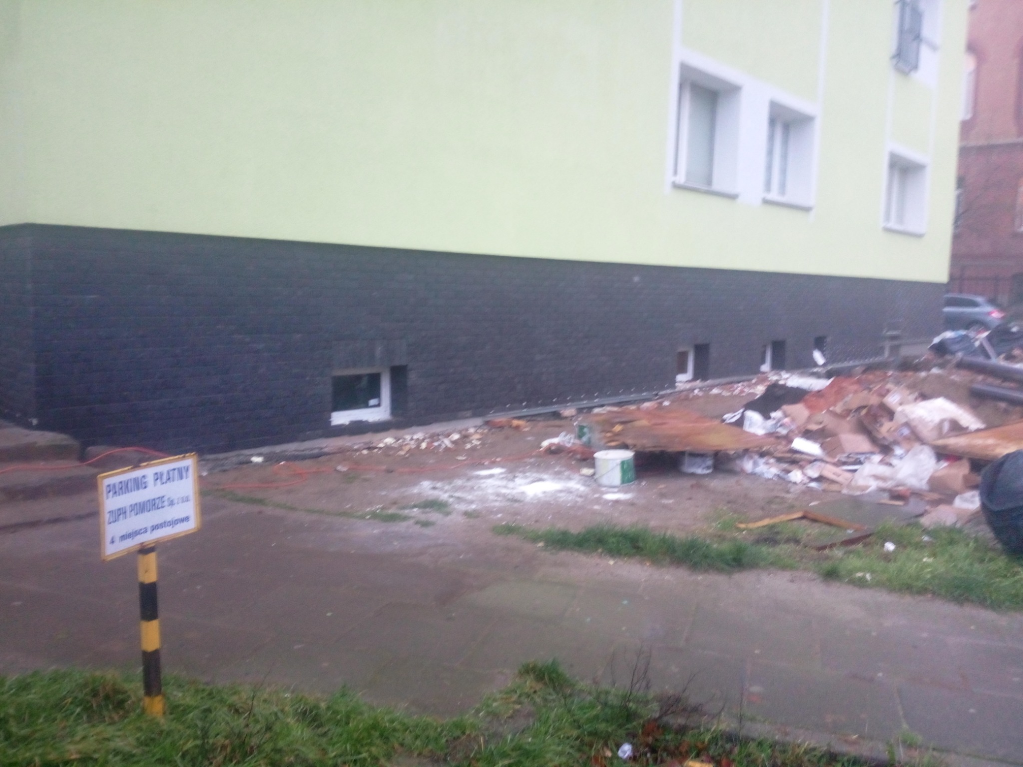 Elewacja budynku z wykończonym cokołem z ciemnej cegły klinkierowej, widoczny bałagan po pracach budowlanych, tablica informacyjna parkingu w pierwszym planie.