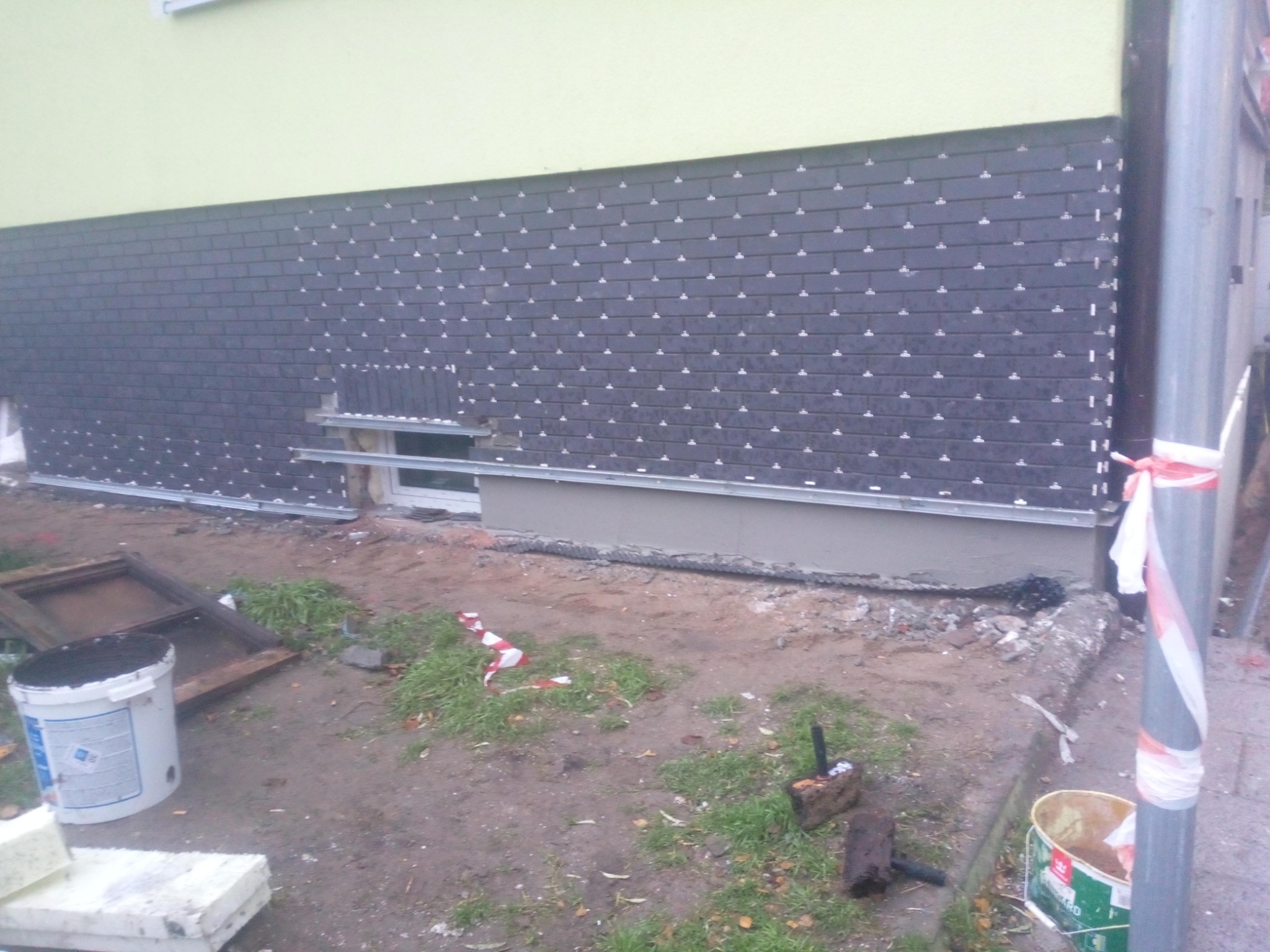 Montaż elewacji z płytek klinkierowych na budynku z oknem, widoczne elementy mocujące i fragmenty rusztowania, teren wokół w trakcie prac ziemnych.