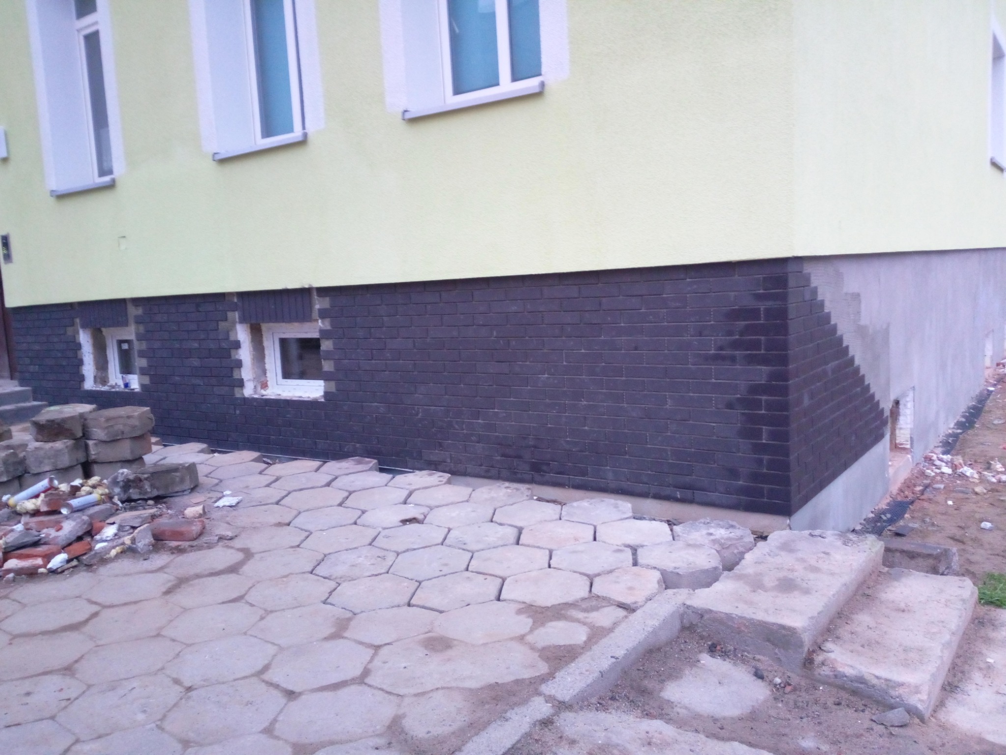 Wykończenie elewacji budynku w Szczecinie z użyciem ciemnej cegły klinkierowej na cokole, widoczne okna piwniczne i fragment chodnika z kostki brukowej, pozostałości materiałów budowlanych obok.