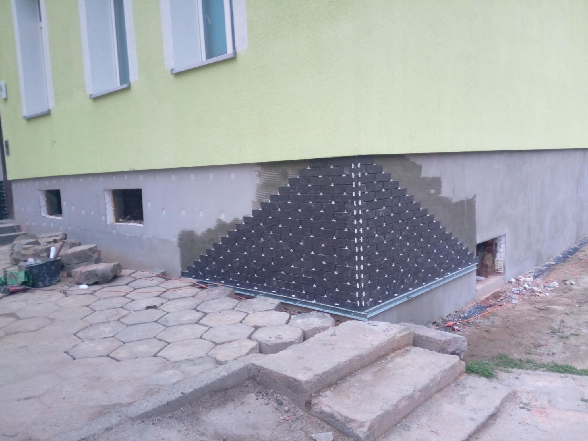 Fragment elewacji budynku w trakcie wykańczania z dekoracyjną okładziną z płytek imitujących cegłę, ułożonych w trójkątny wzór, widoczne elementy mocujące, na pierwszym planie betonowe schody...