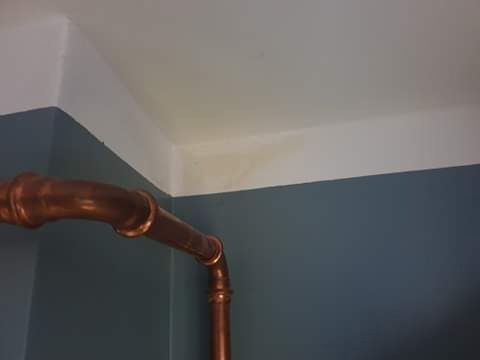 Miedziana rura instalacji wodnej, zamontowana w rogu pomieszczenia z pomalowanymi ścianami na kolor niebieski i białym sufitem z widocznym odbarwieniem.