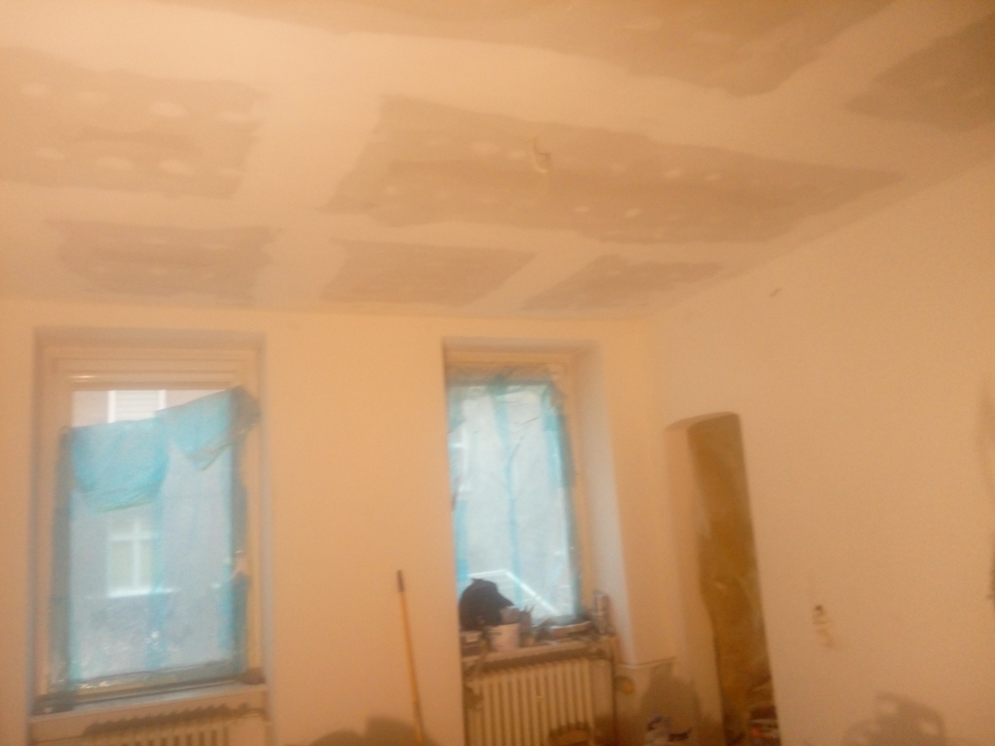 Pokój w trakcie prac remontowych, zabezpieczone okna folią, widoczne fragmenty sufitu z nałożoną warstwą szpachli, przygotowane do dalszej obróbki.