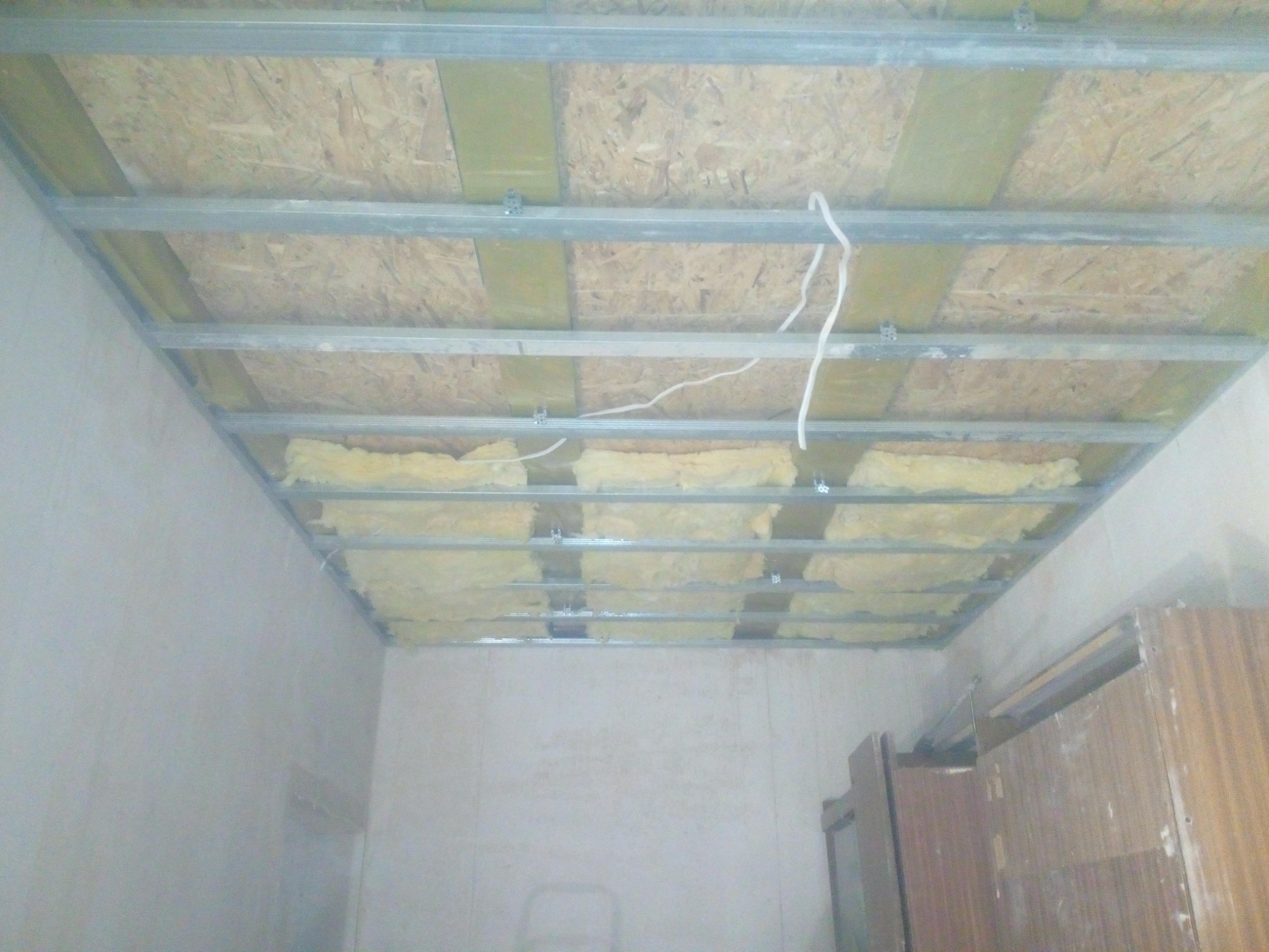 Montaż izolacji z wełny mineralnej w metalowym stelażu sufitu podwieszanego, widoczne przewody elektryczne i płyty OSB.