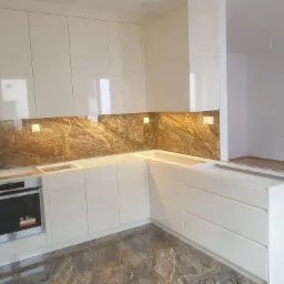 Jasna, lakierowana zabudowa kuchenna w trakcie montażu, z marmurowym panelem nad blatem i wbudowanym piekarnikiem, widok na puste pomieszczenie w tle.