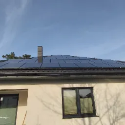 Panele słoneczne na dachu domu jednorodzinnego z kominem, widoczne z dołu, z fragmentem drzewa iglastego w kadrze.