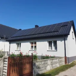 Dom jednorodzinny z panelami słonecznymi na dachu, widok z poziomu ulicy, ogrodzenie betonowe i czerwone kwiaty w ogródku.