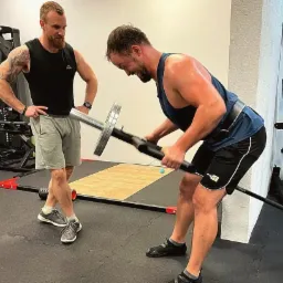 Mężczyzna w koszulce na ramiączkach ćwiczy podnoszenie ciężarów z pomocą instruktora w siłowni. Instruktor z brodą i tatuażem na ramieniu obserwuje postępy.