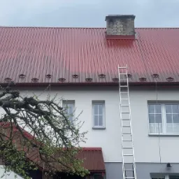 Częściowo umyty, czerwony dach blaszany z widocznym, ciemniejszym nalotem i metalową drabiną opartą o ścianę budynku, komin z cegły na dachu.