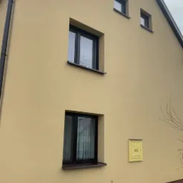 Żółta elewacja domu z trzema oknami o czarnych ramach, skrzynka gazowa, widoczny fragment dachu i rynny, na trawniku worek z zaprawą murarską.