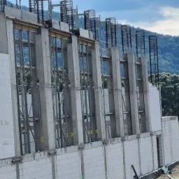 Wznoszenie ściany z betonu komórkowego Ytong z użyciem szalunków i zbrojenia pionowego, widoczny pracownik w żółtym kasku i kamizelce, w tle zalesione wzgórza.