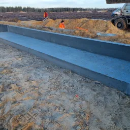 Betonowy fundament w trakcie budowy, z widocznymi pracownikami w odblaskowych kamizelkach i sprzętem budowlanym w tle, na piaszczystym terenie.